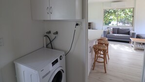 A laundry area with a white washing machine, a ceiling fan, and a view of a living room with a sofa and a table. - Cedric Cottage, 1B Cedric Court (Mallacoota)