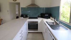 A modern kitchen with white cabinetry, a built-in oven, a microwave, and a large window providing a view of greenery outside. - Cedric Cottage, 1B Cedric Court (Mallacoota)