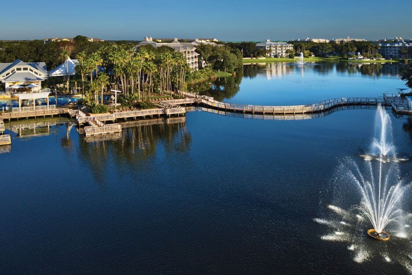 A lake with a fountain, surrounded by buildings and palm trees.