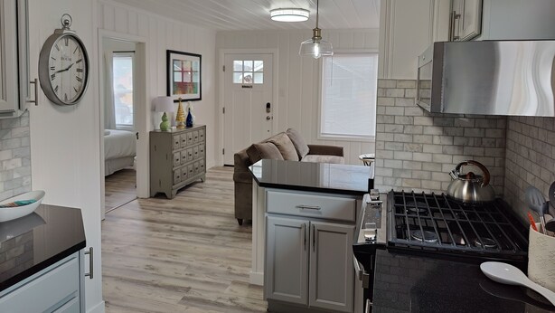 A modern kitchen with a stove, sink, and cabinets. There is a dining area with a couch and a clock on the wall.