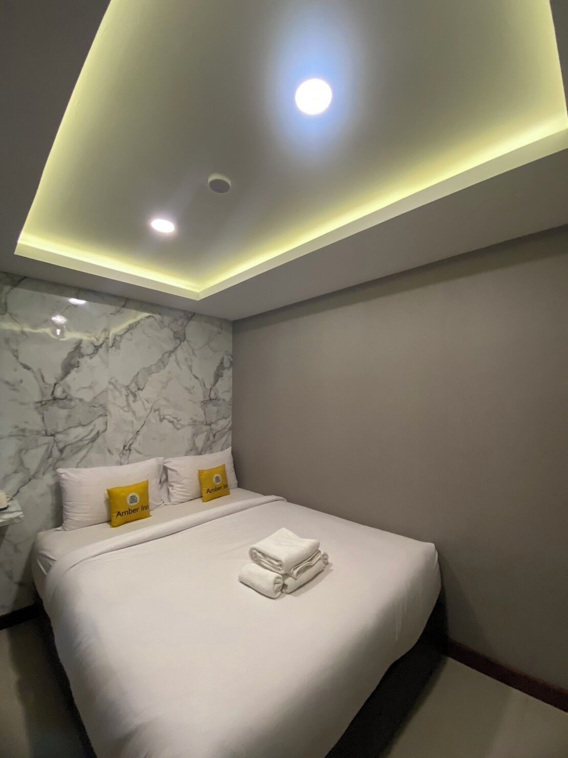 A modern hotel room with a marble wall, a neatly made bed with white linens, and a ceiling with recessed lighting.