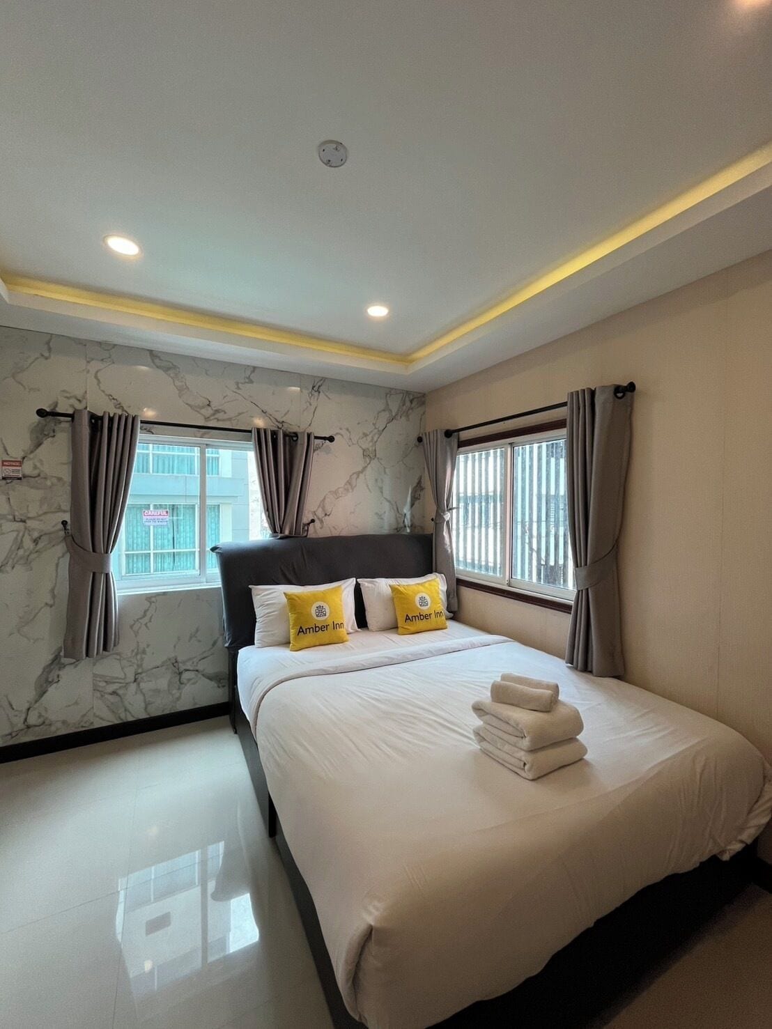 A hotel room with a bed, two pillows, a headboard, a window with curtains, a ceiling light, and a marble wall.