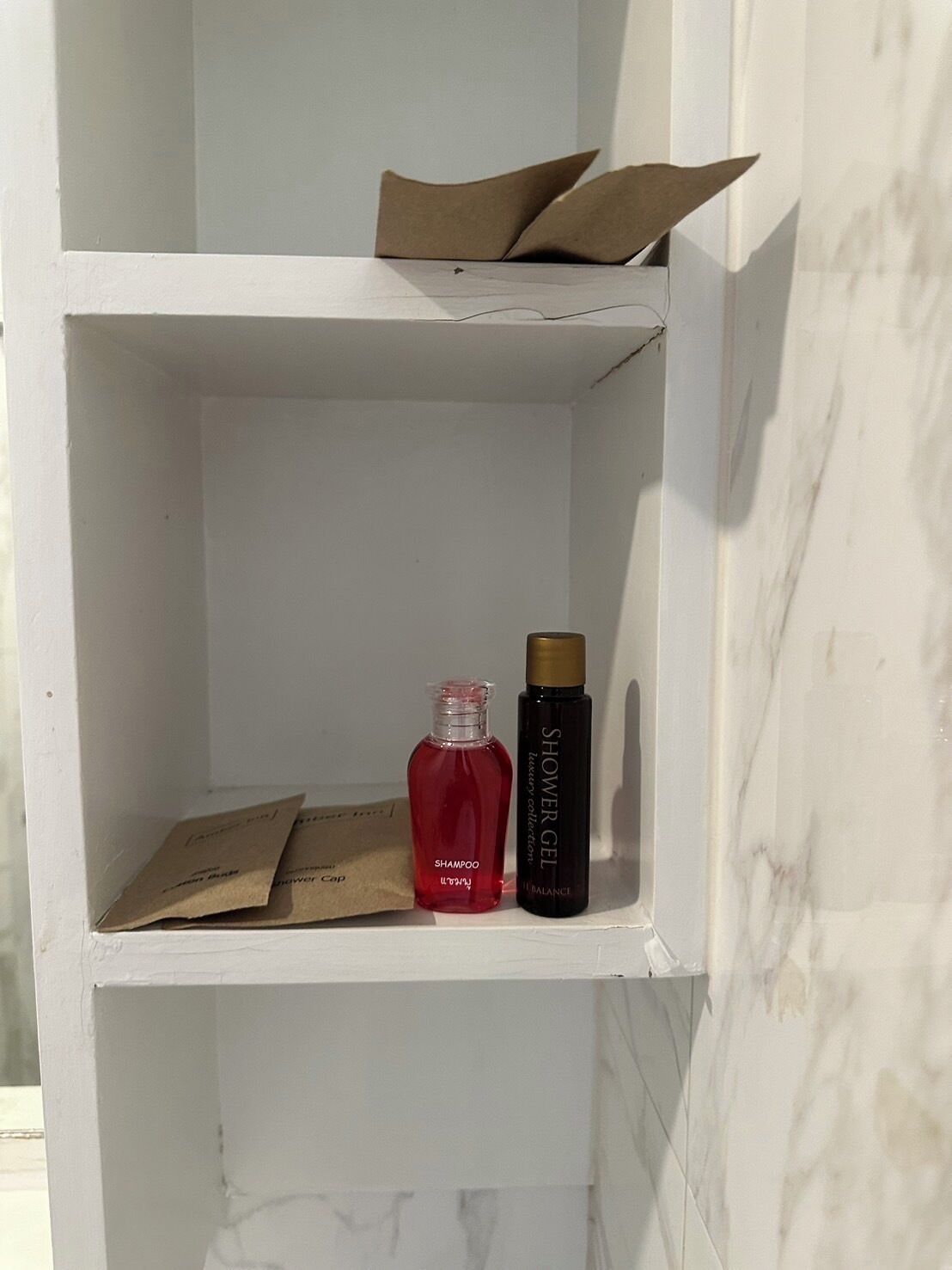 A white shelf with a marble wall background, containing a red bottle labeled 'SHAMPOO', a black bottle labeled 'SHOWER GEL', and two brown paper bags.