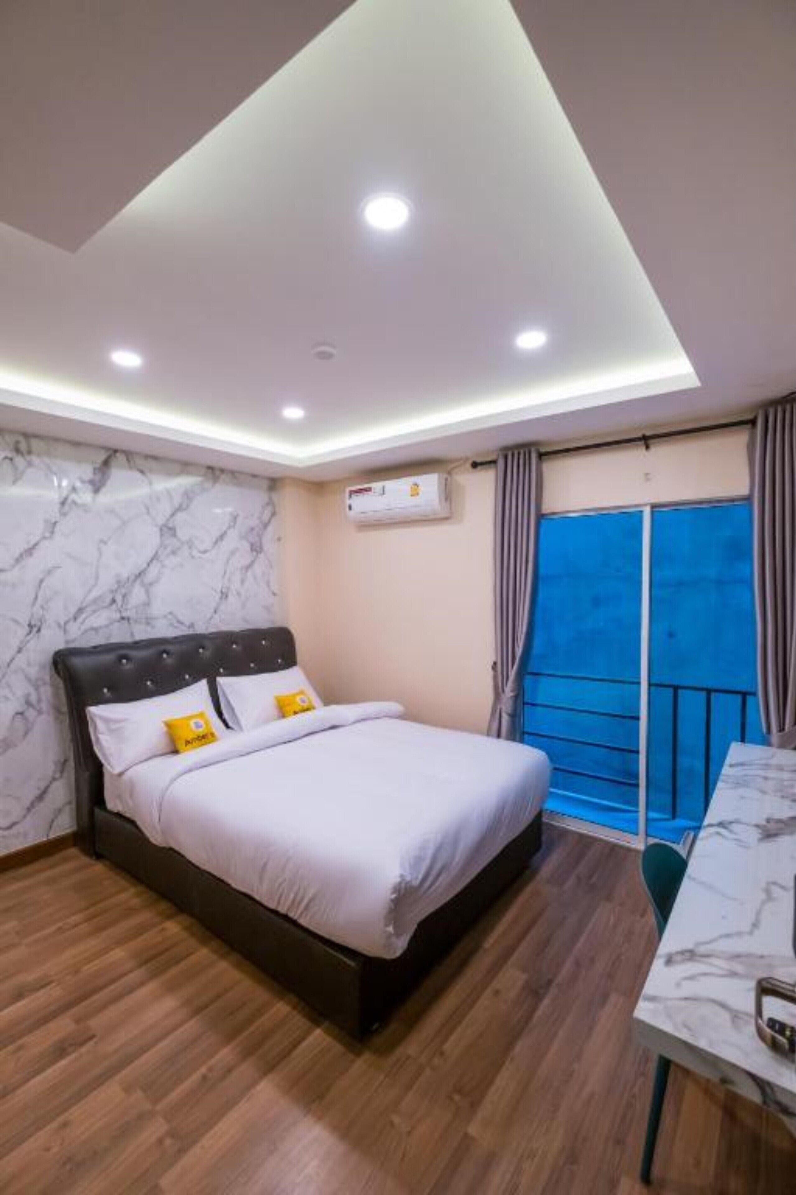 A modern bedroom with a bed, wooden flooring, marble wall, ceiling lights, and a balcony with curtains.