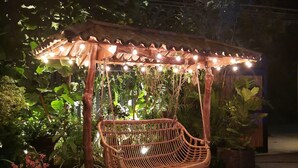 Una mecedora de madera bajo una pérgola con luces colgantes, rodeada de plantas y grava.