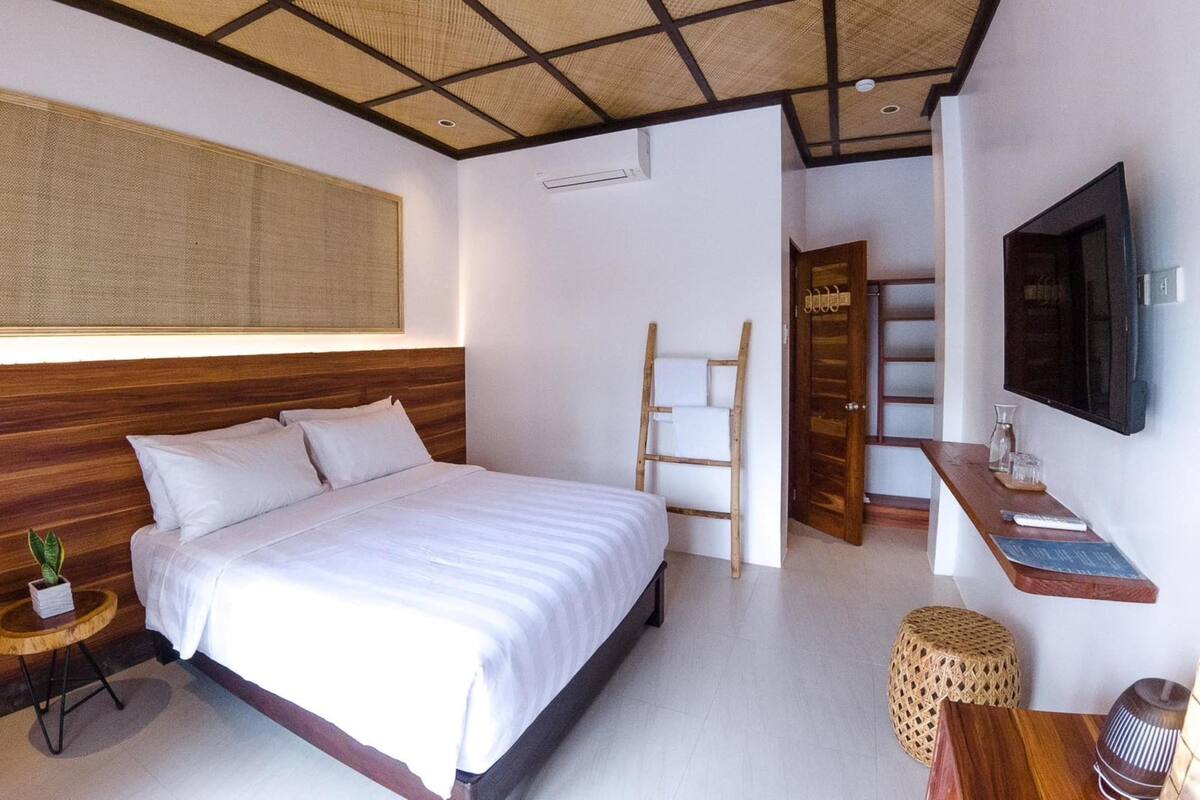 Un dormitorio moderno con una cama grande, cabecera de madera, televisión montada en la pared y una pequeña mesa de madera con una planta.