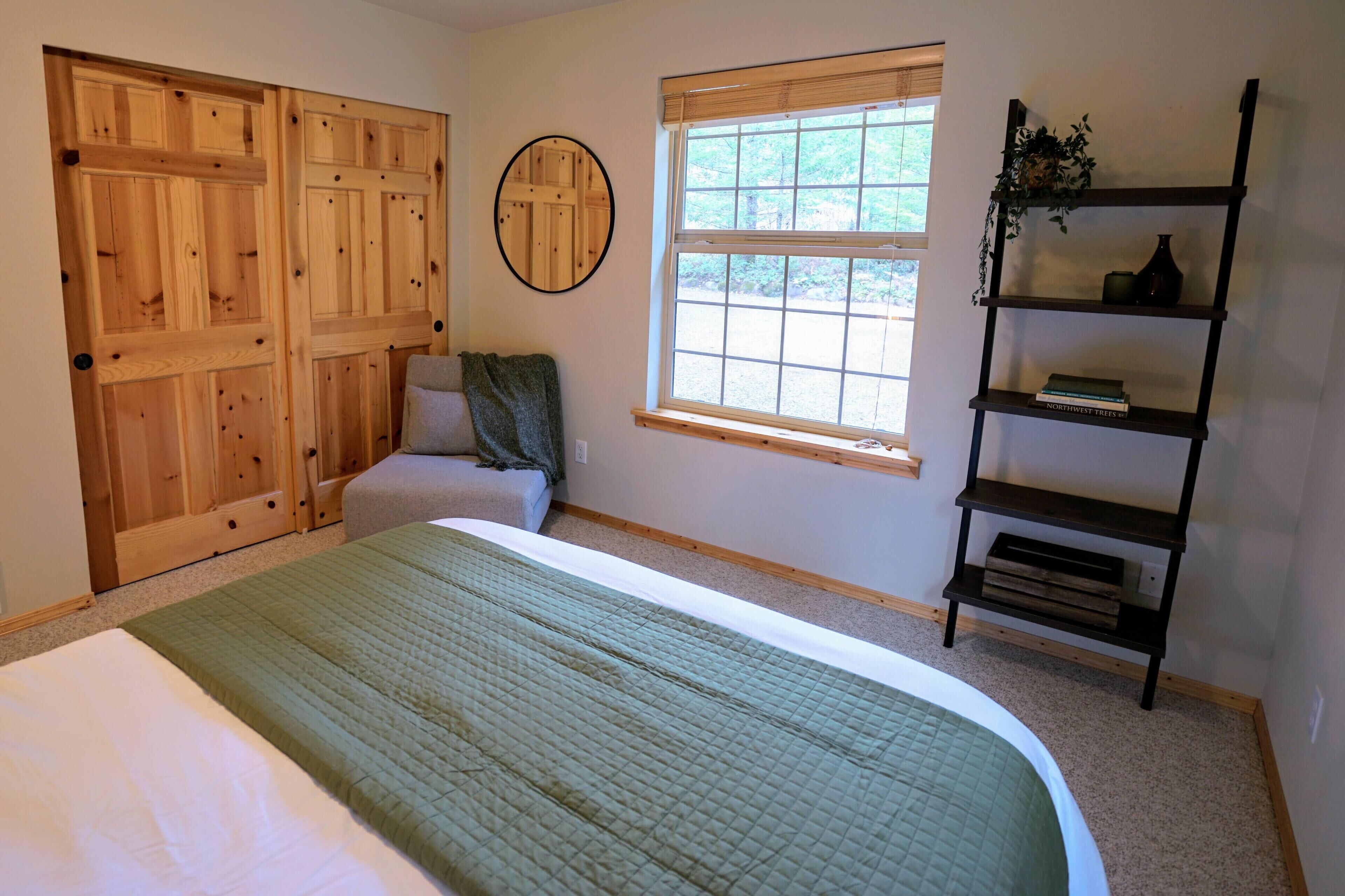 A bedroom with a large bed, a wooden wardrobe, a window with blinds, and a bookshelf.