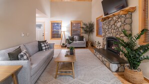 Un salon avec une cheminée en pierre, un canapé gris, une table basse en bois et un téléviseur fixé au mur.