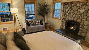 Une chambre à coucher avec un grand lit, un canapé, une cheminée en pierre et une fenêtre donnant sur la forêt.