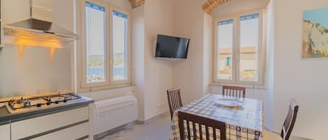 A kitchen with a gas stove, a built-in oven, and a TV mounted on the wall. There are two windows with shutters, a table with chairs, and a painting on the wall.