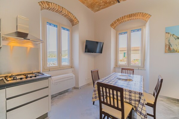 A kitchen with a gas stove, a built-in oven, and a TV mounted on the wall. There are two windows with shutters, a table with chairs, and a painting on the wall.