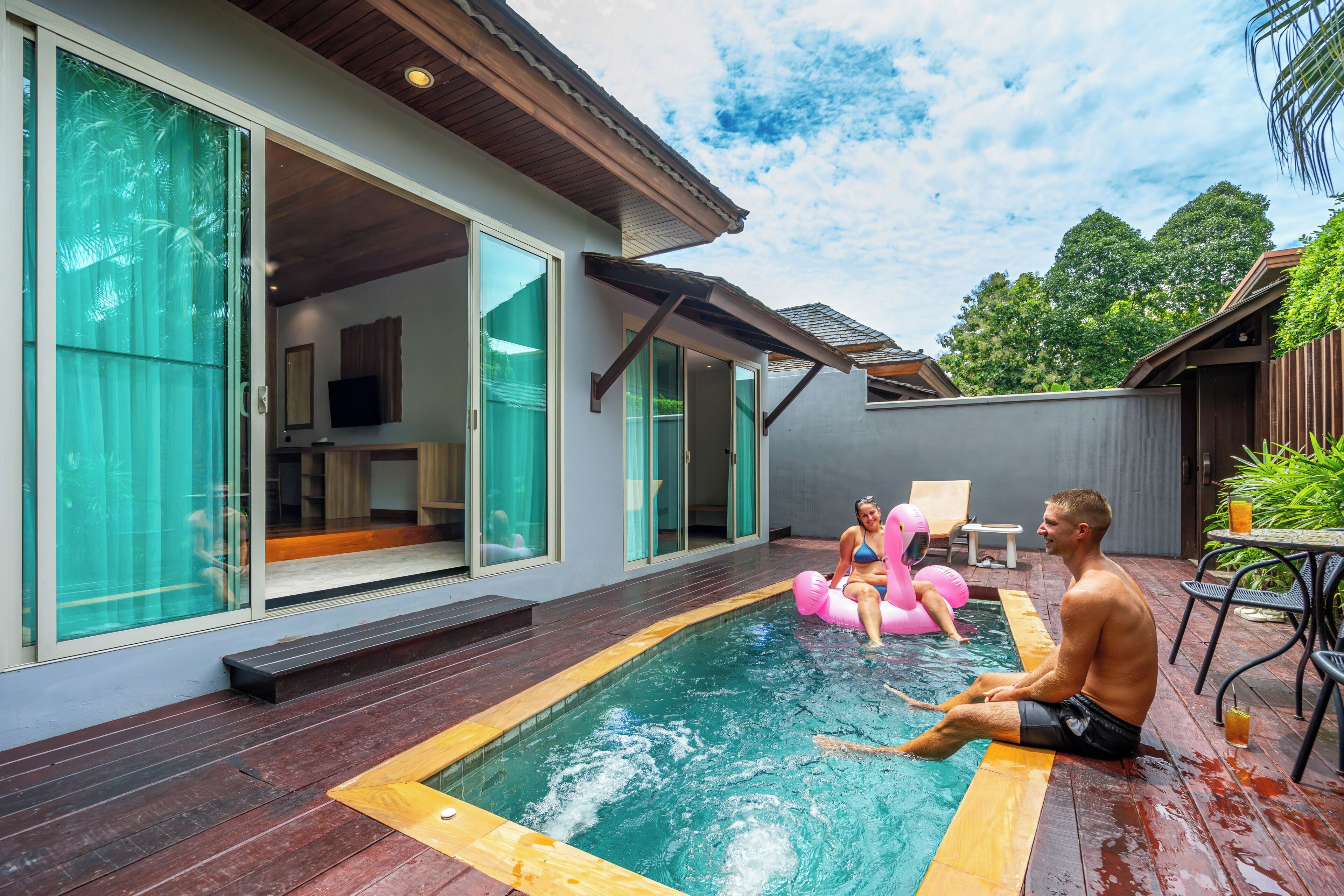 A swimming pool with wooden decking, a glass door entrance, and a patio area with outdoor seating.