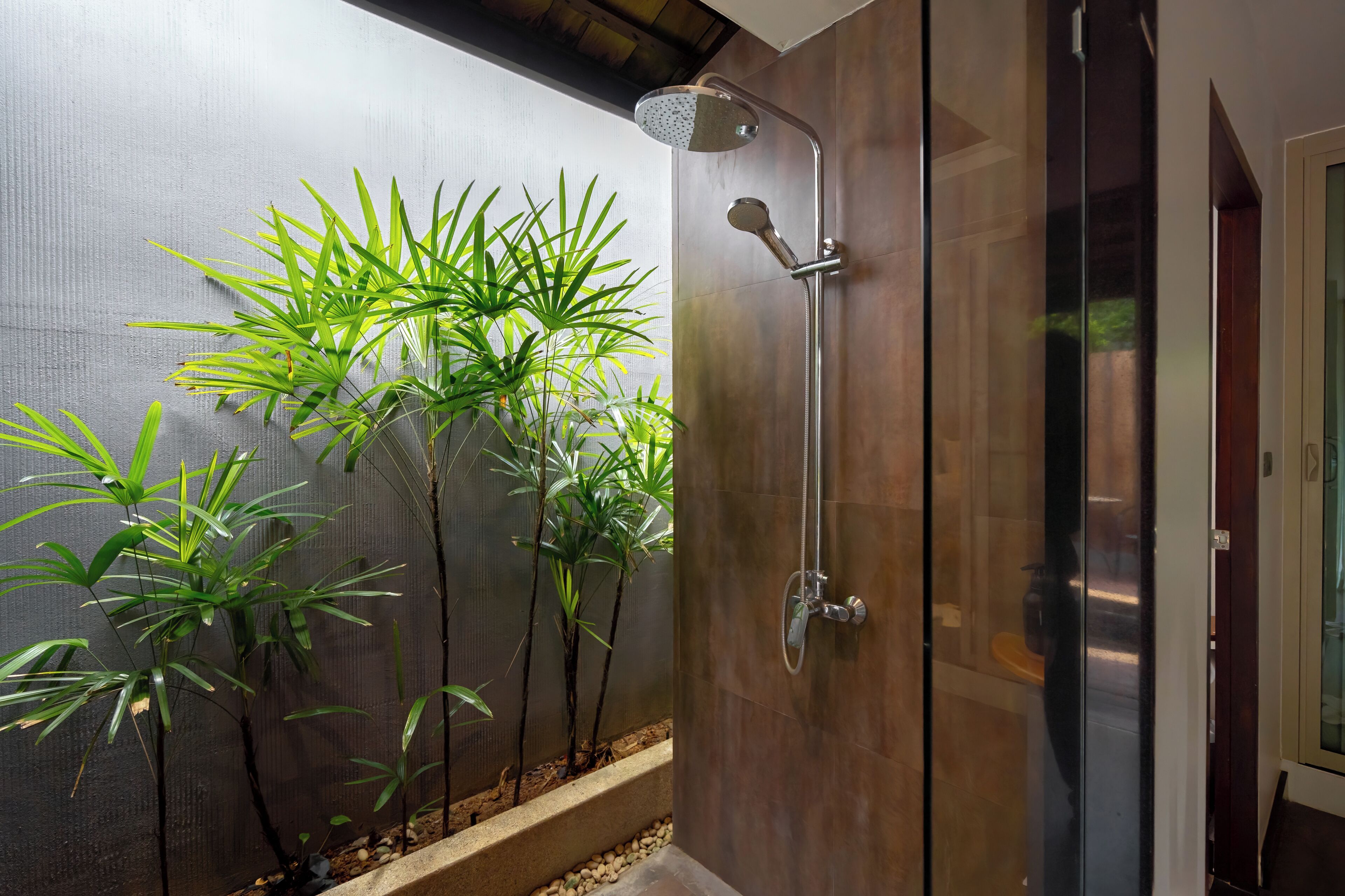 A modern bathroom with a glass shower enclosure, a wooden bench, and a row of green plants along the wall.