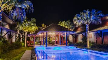 Vista nocturna de un complejo turístico con piscina, palmeras y una zona de estar cubierta.