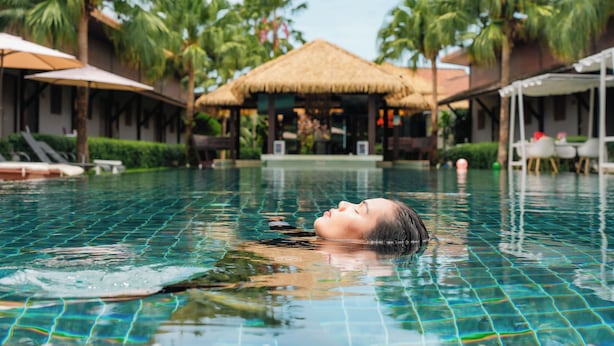 Una piscina con una persona che si rilassa, circondata da palme e costruzioni con tetti di paglia.