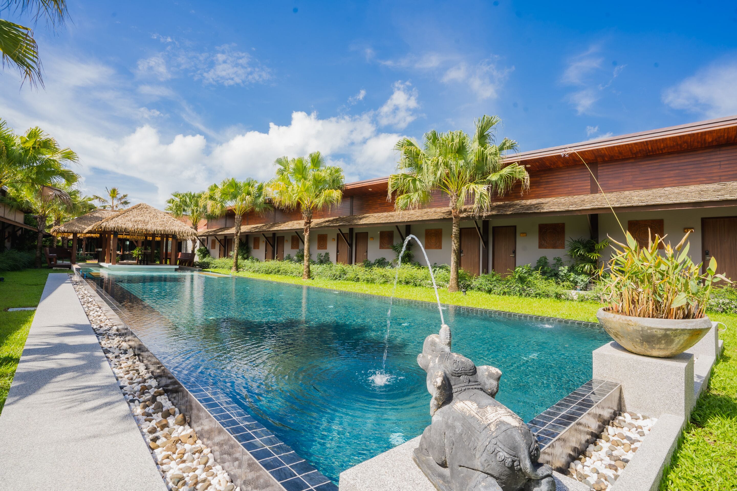 Una piscina con una escultura de piedra, rodeada de palmeras y un edificio con techo de paja.