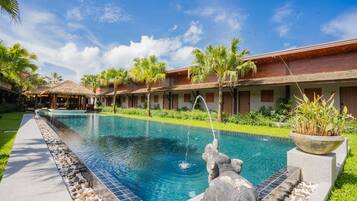 Una piscina con una escultura de piedra, rodeada de palmeras y un edificio con techo de paja.