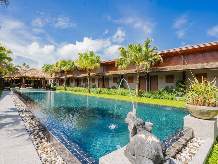 Una piscina con una escultura de piedra, rodeada de palmeras y un edificio con techo de paja.