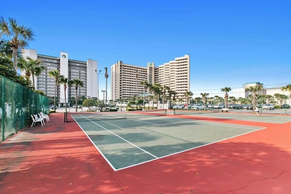 An outdoor tennis court with a red surface, surrounded by a green fence, and adjacent to a row of tall buildings.