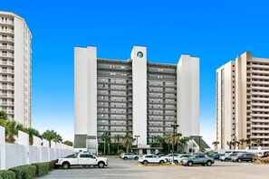 A modern hotel with a central tower and multiple smaller buildings. It features a parking lot with several cars and a white fence along the perimeter.