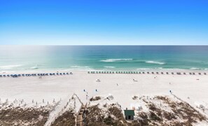 A beach with rows of umbrellas and a few people. There is a small building and some vegetation in the foreground.