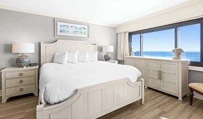 A hotel room with a large bed, a nightstand with a lamp, a dresser, and a window with a view of the ocean.