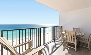 A balcony with a view of the beach and ocean, wooden chairs, and a table.
