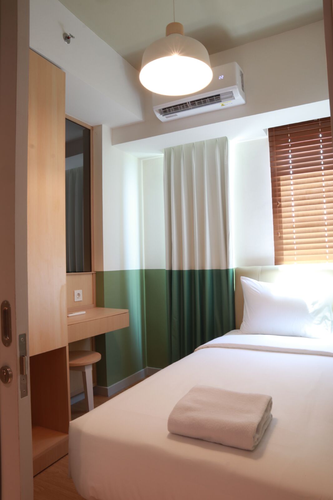 A hotel room with a bed, a bedside table, a mirror, an air conditioning unit, and a window with blinds.