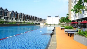 A swimming pool with clear blue tiles, surrounded by lounge chairs and umbrellas, with modern buildings in the background. - Hotel Osaka Pik2 (Kosambi)