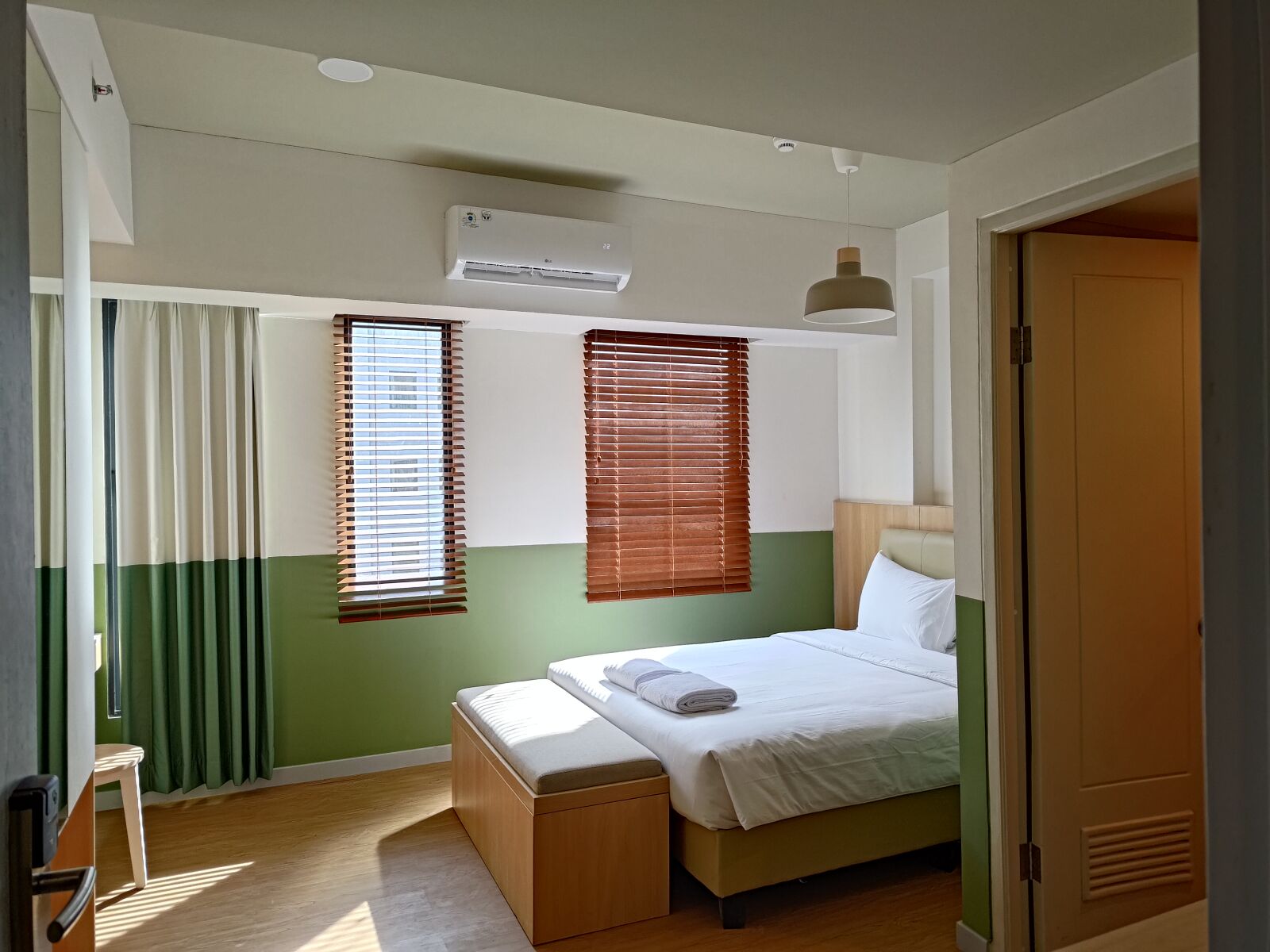 Ein Schlafzimmer mit Bett, Nachttisch, Fenster mit Jalousien, Klimaanlage und einer Pendelleuchte.