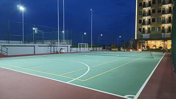 An outdoor tennis court at night with floodlights, surrounded by a fence and a building with balconies.