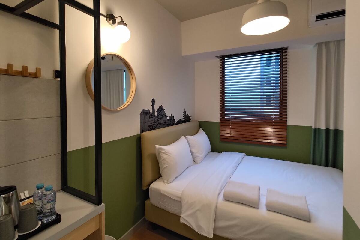 A hotel room with a bed, a round mirror, a cityscape artwork, a window with blinds, and a hanging light fixture.