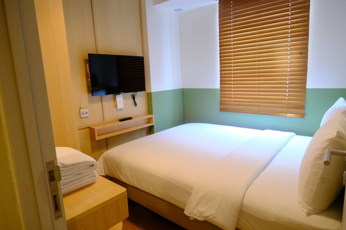 A hotel room with a bed, a TV mounted on the wall, a window with blinds, and a small table with a towel.