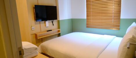 Une chambre d’hôtel avec un lit, un téléviseur fixé au mur, une fenêtre avec des stores et une petite table sur laquelle est posée une serviette.