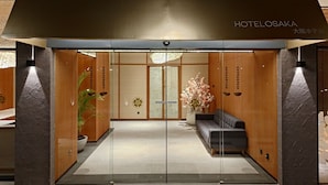 A hotel entrance with a modern design, featuring a reception desk, a seating area, and a large potted plant. - Hotel Osaka Pik2 (Kosambi)