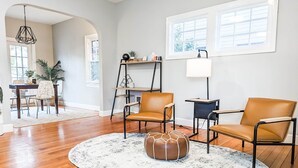 Ein modernes Wohnzimmer mit zwei Ledersesseln, einem runden Hocker, einem weißen Teppich und einem Holztisch. Im Hintergrund ist ein Essbereich mit Tisch und Stühlen zu sehen.
