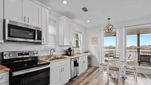 A modern kitchen with white cabinets, a stainless steel microwave, and a black stove. There is a dining area with white chairs and a table, and a large window offering a view of the city. - Waterview Villa~Steps to Beach~Pool~King Bed (Destin)