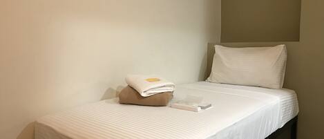 Una cama individual con un colchón blanco y una almohada color beige, una toalla doblada y un control remoto sobre la cama.