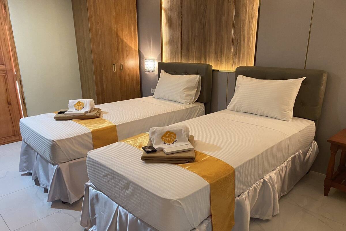 Une chambre d’hôtel avec deux lits simples, chacun recouvert d’une courtepointe blanche agrémentée d’un motif doré. On y trouve des armoires en bois et une petite table en bois.
