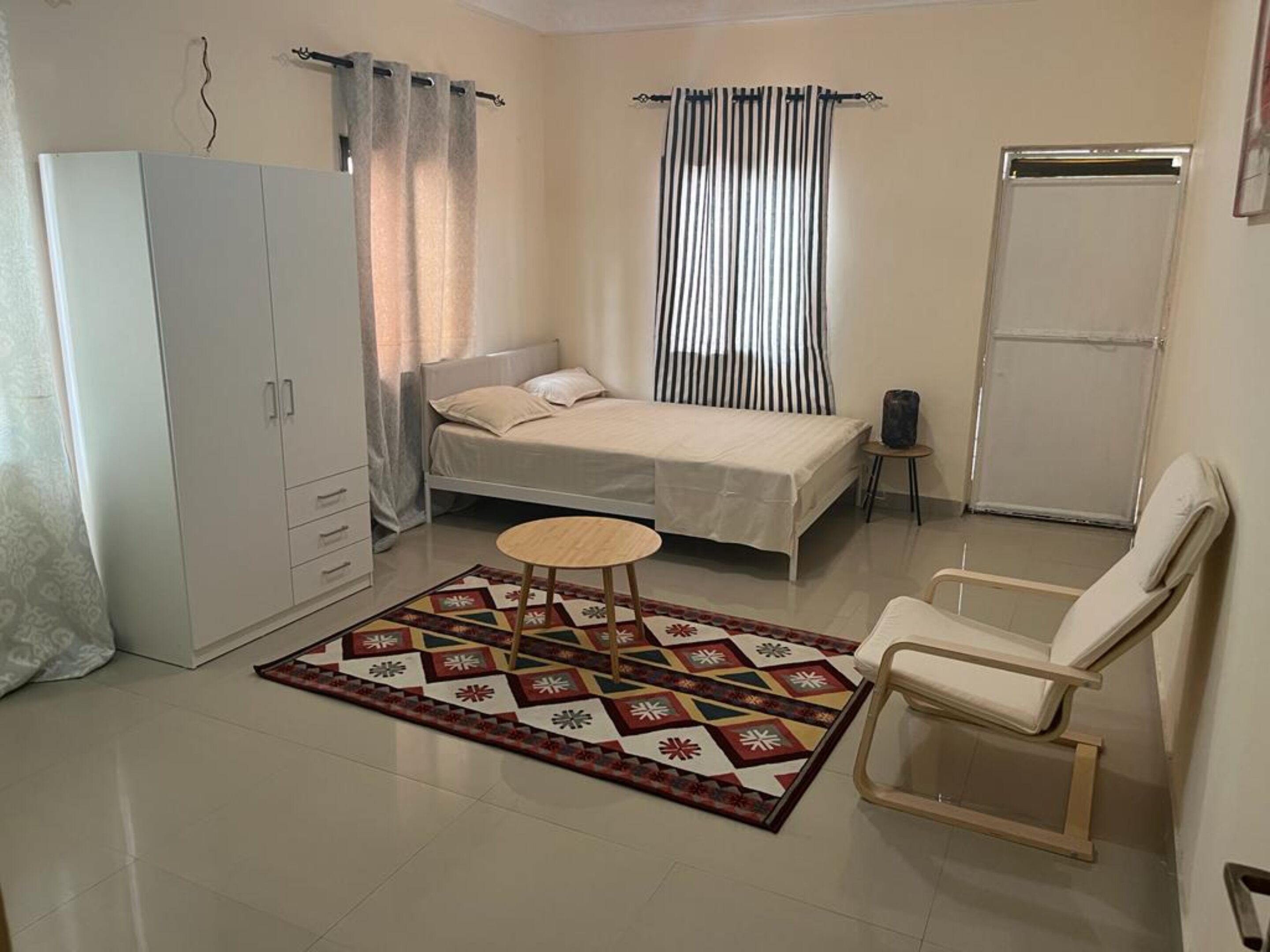 Quarto pequeno e bem iluminado com uma cama, uma cadeira, um guarda-roupa e uma mesinha.