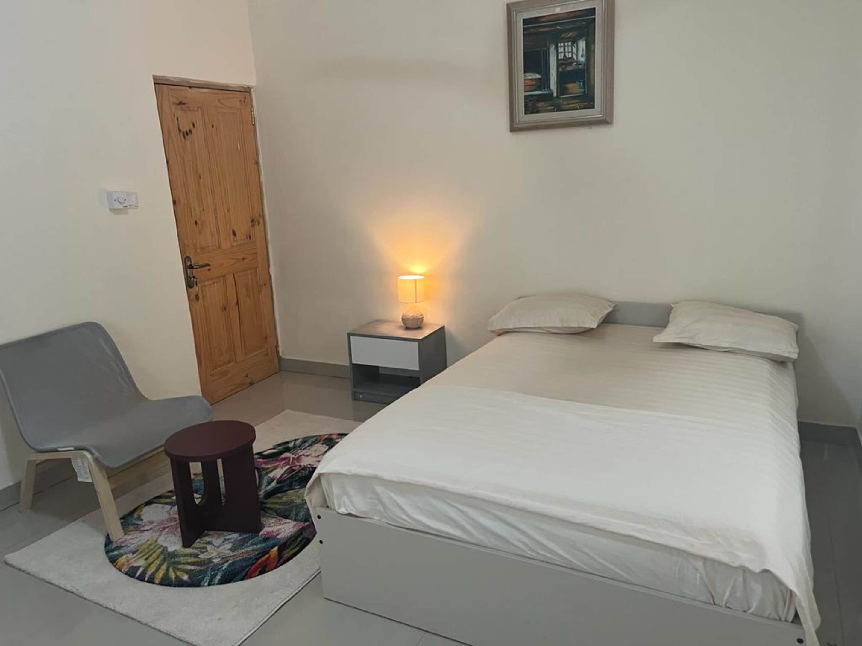 A bedroom with a bed, a chair, a nightstand with a lamp, a door, and a framed picture on the wall.