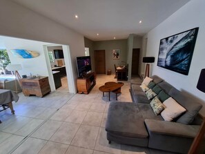 Un grand espace de vie comprenant un canapé, une table basse et un téléviseur. On y trouve également un coin repas avec des chaises et une table, ainsi qu'une cuisine visible à travers une porte ouverte.