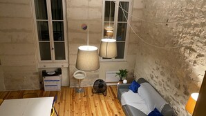 Ein gemütlicher Wohnraum mit Holzboden, einem grauen Sofa mit blauen Kissen, einem weißen Tisch und einem kleinen runden Beistelltisch. Zwei Fenster mit Vorhängen, eine Wandlampe und eine Heizung sind vorhanden.