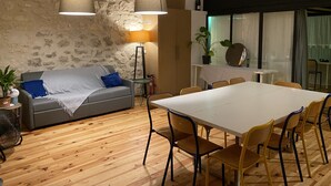 Ein modernes Wohnzimmer mit Steinwand, einem grauen Sofa, einem Esstisch mit Stühlen und einem großen Fenster mit Vorhängen.