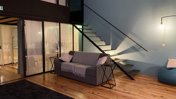 Ein modernes Wohnzimmer mit einem grauen Sofa, einem blauen Sitzsack und Holzdielen. Eine Treppe mit schwarzer Geländer führt auf eine obere Ebene.