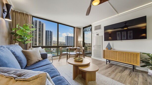A modern living room with a blue sofa, a round wooden coffee table, and a view of tall buildings through large windows.