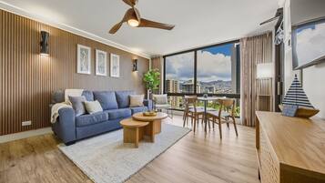 Un salon moderne avec un canapé bleu, un plancher en bois, un ventilateur de plafond et de grandes fenêtres donnant sur la ville.
