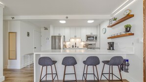 A modern kitchen with white cabinets, a stainless steel refrigerator, a microwave, and a built-in oven. There are three bar stools in front of the counter, and a clock on the wall.