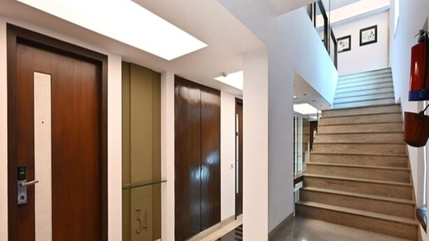 Un couloir d’hôtel moderne, avec des portes en bois, un ascenseur en verre et un escalier menant à un étage supérieur.