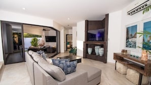 Una sala de estar moderna con un sofá gris, una mesa de centro y un televisor montado en la pared. Se ve una estantería con objetos decorativos y una planta en maceta.